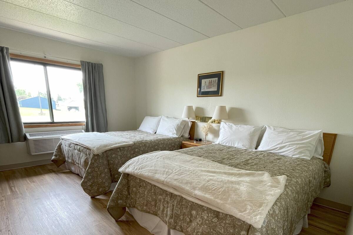 A hotel room with two beds, a large window with curtains, a picture on the wall, and a wooden floor.