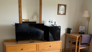 Habitación de hotel con tocador de madera, televisor, refrigerador, escritorio y silla, y una lámpara.