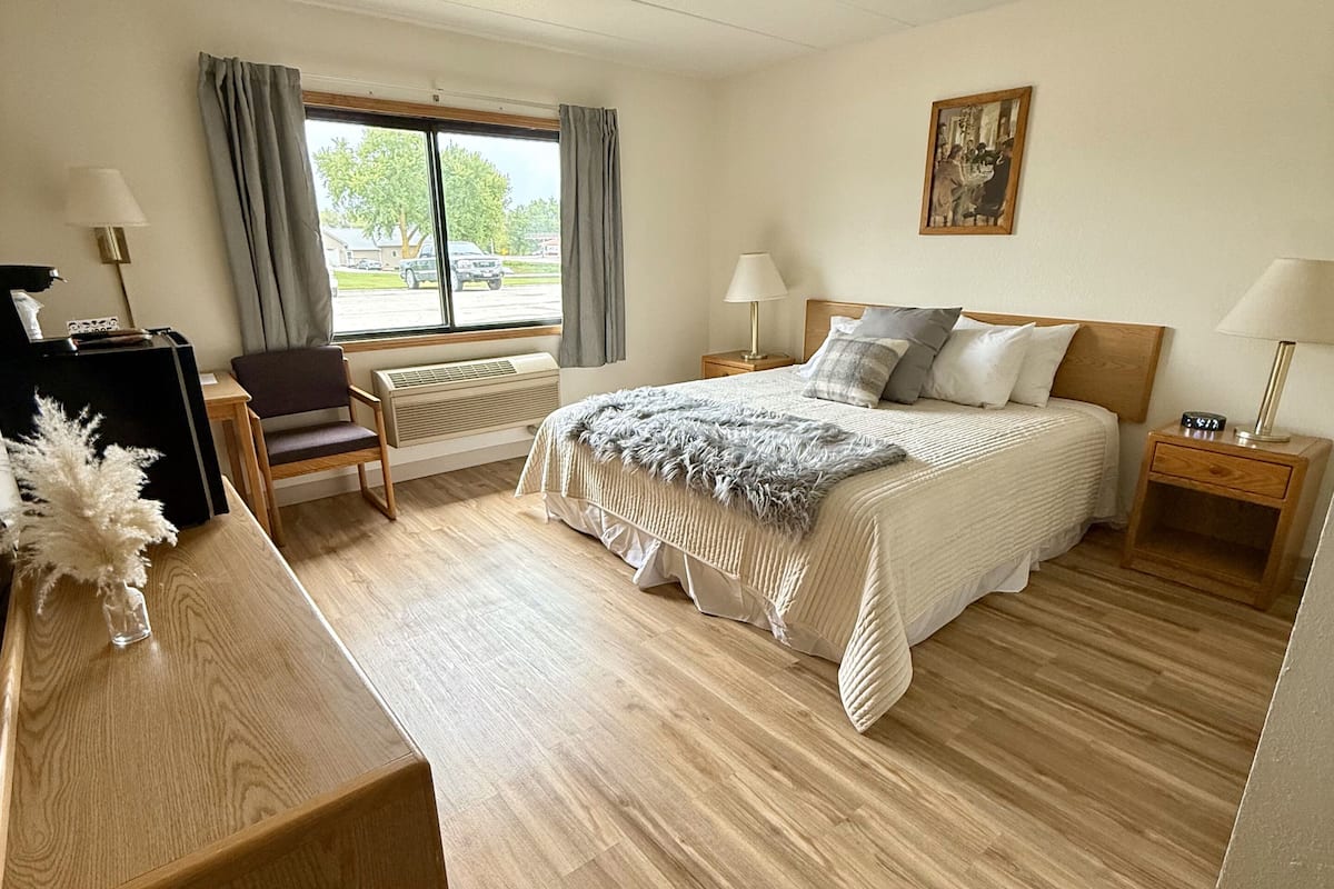 A hotel room with a bed, bedside tables, a desk, a chair, a window with curtains, and a painting on the wall.