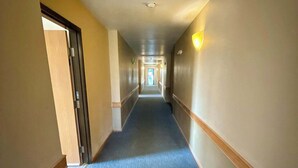Un largo y estrecho pasillo de hotel con alfombra azul, detalles de madera y una lámpara en la pared derecha.