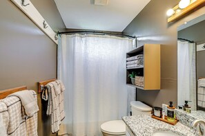 Baño con inodoro, lavamanos y cortina de ducha. Hay toallas colgadas en un perchero y un estante con artículos de aseo.