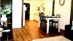 Un comedor con una mesa y sillas de madera, un reloj en la pared y una cocina al fondo.
