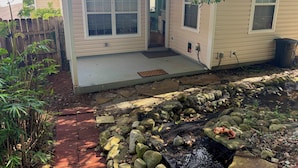 Een huis met een veranda, een kleine tuin met een waterpartij en een houten omheining.
