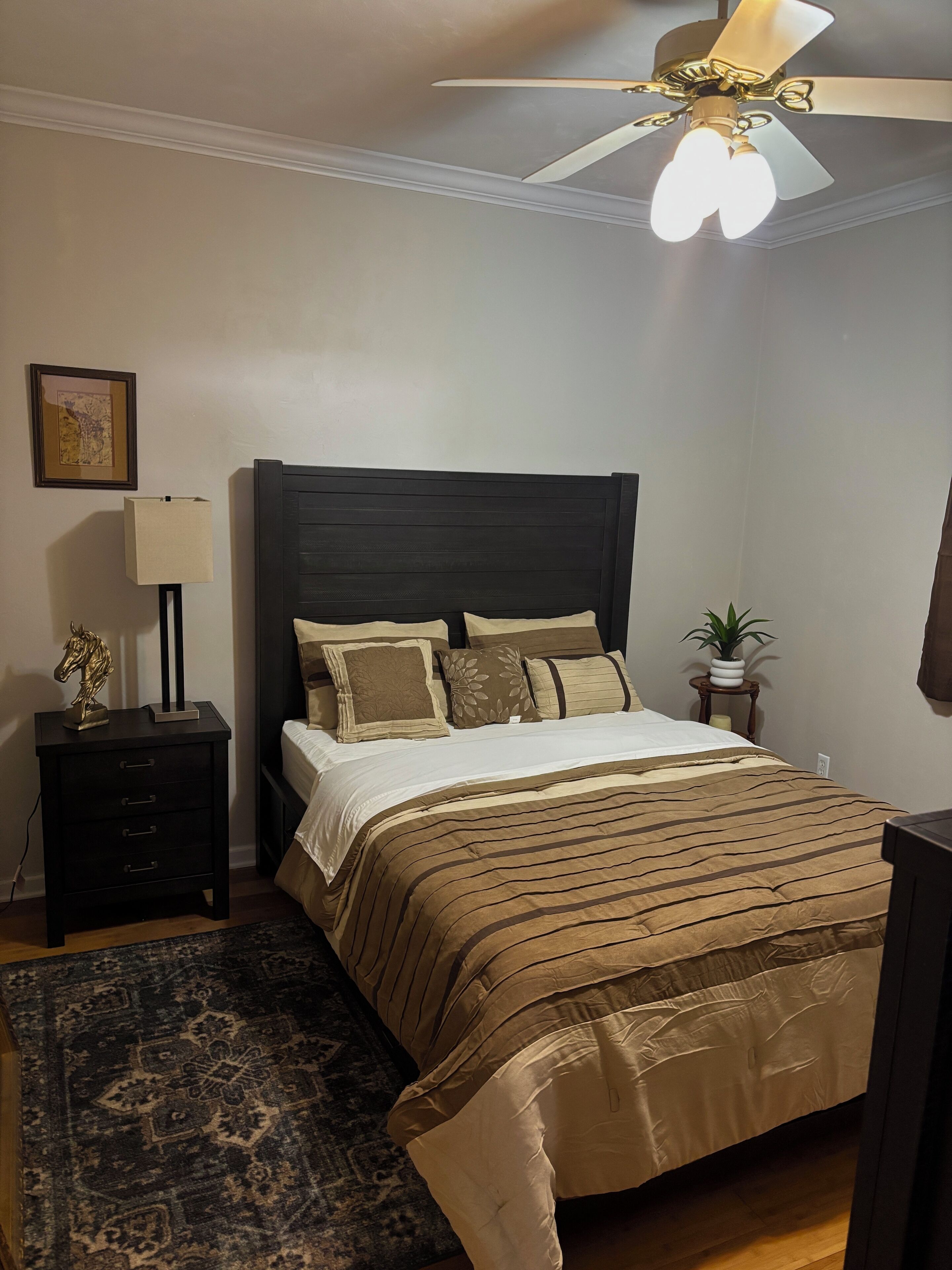 A bedroom with a dark wooden headboard, a patterned bedspread, a nightstand with a lamp, a framed picture on the wall, and a ceiling fan.