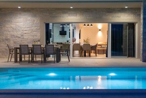 An outdoor pool area with a stone wall, a dining area with chairs and tables, and a sliding glass door leading to an interior space. - Villa FUSKA with private pool (Pinezići)
