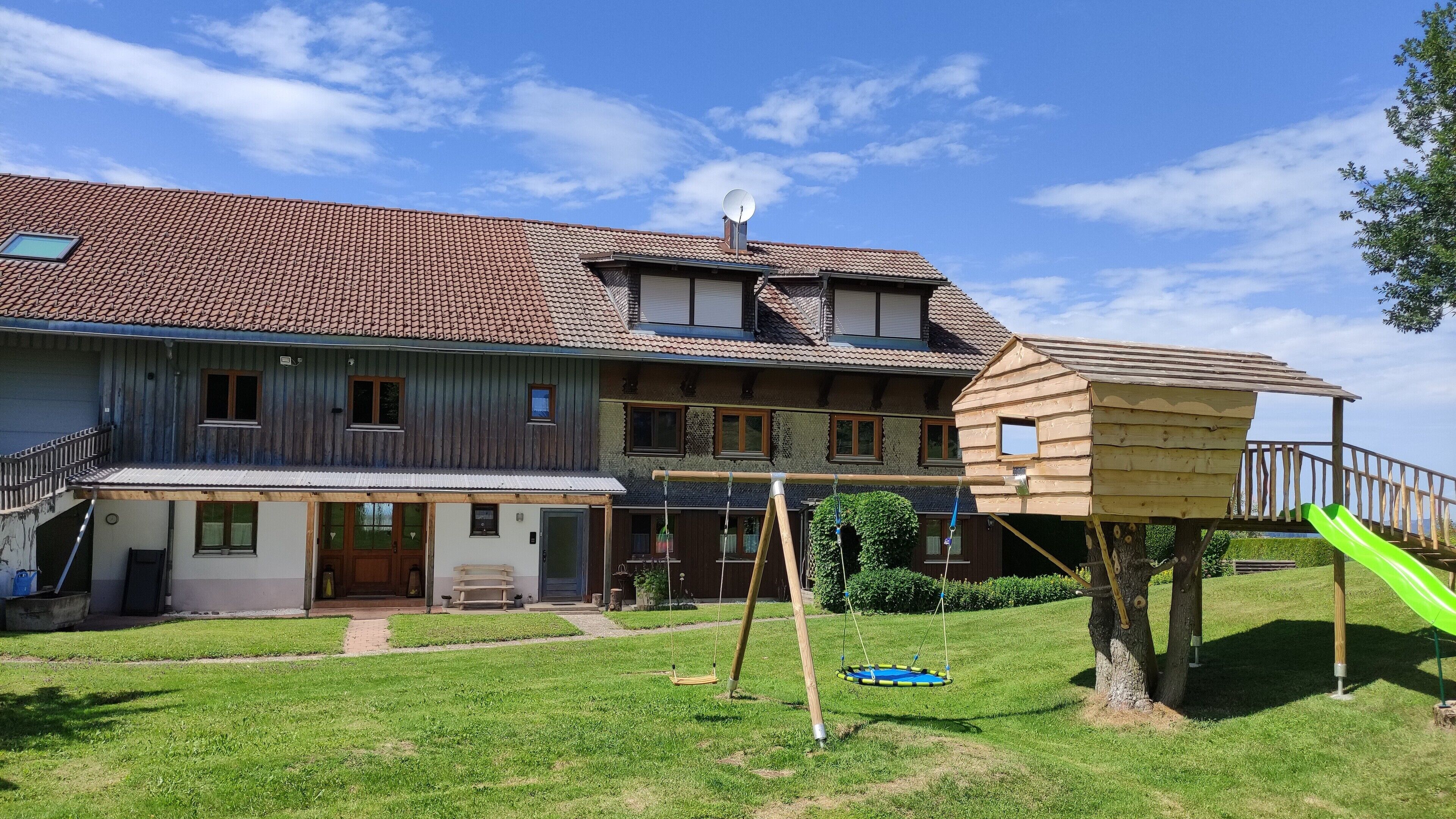 A two-story house with a balcony, a wooden playhouse with a slide, and a green lawn.