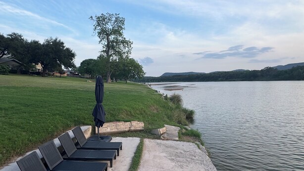 Un patio junto al lago con sillones de descanso y una sombrilla, un área verde y vistas al lago y colinas lejanas.
