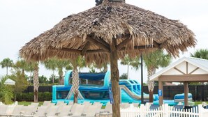 Une plage avec des parasols en chaume, des chaises longues et un toboggan aquatique en arrière-plan.