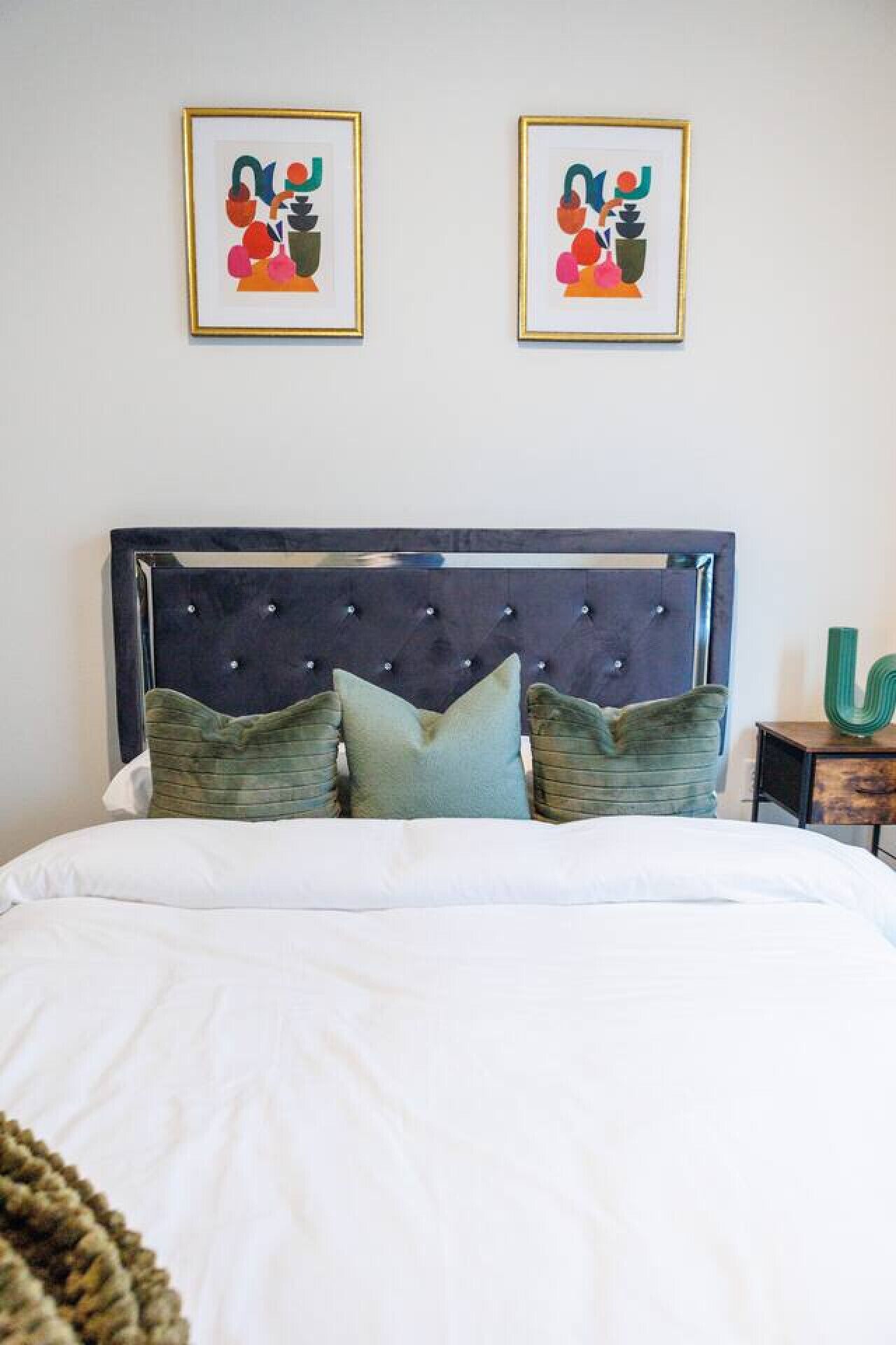 Ein Schlafzimmer mit einer gesteppten Kopfteil, einem Bett mit weißer Bettwäsche und zwei gerahmten abstrakten Bildern an der Wand.