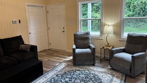 Une pièce avec un canapé de couleur sombre, deux fauteuils, un tapis à motifs et deux fenêtres donnant sur un paysage verdoyant.