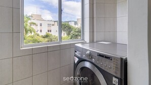 A compact laundry room with a washing machine, a window with a view of buildings and greenery, and tiled walls. - Apt 700m From Ingleses Beach Mab0203 (Florianópolis)