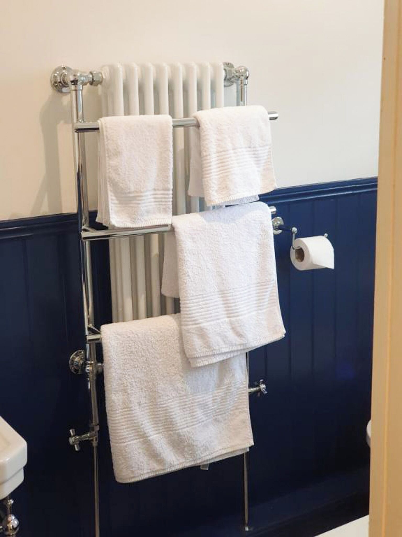 A bathroom with a towel rack holding four white towels, a toilet paper holder, and a radiator.