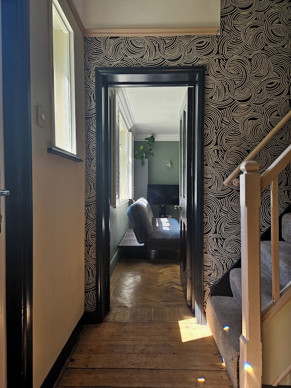A narrow hallway with a wooden floor leading to a room with a bed and a desk. - The Lodge Stanwell (Staines)