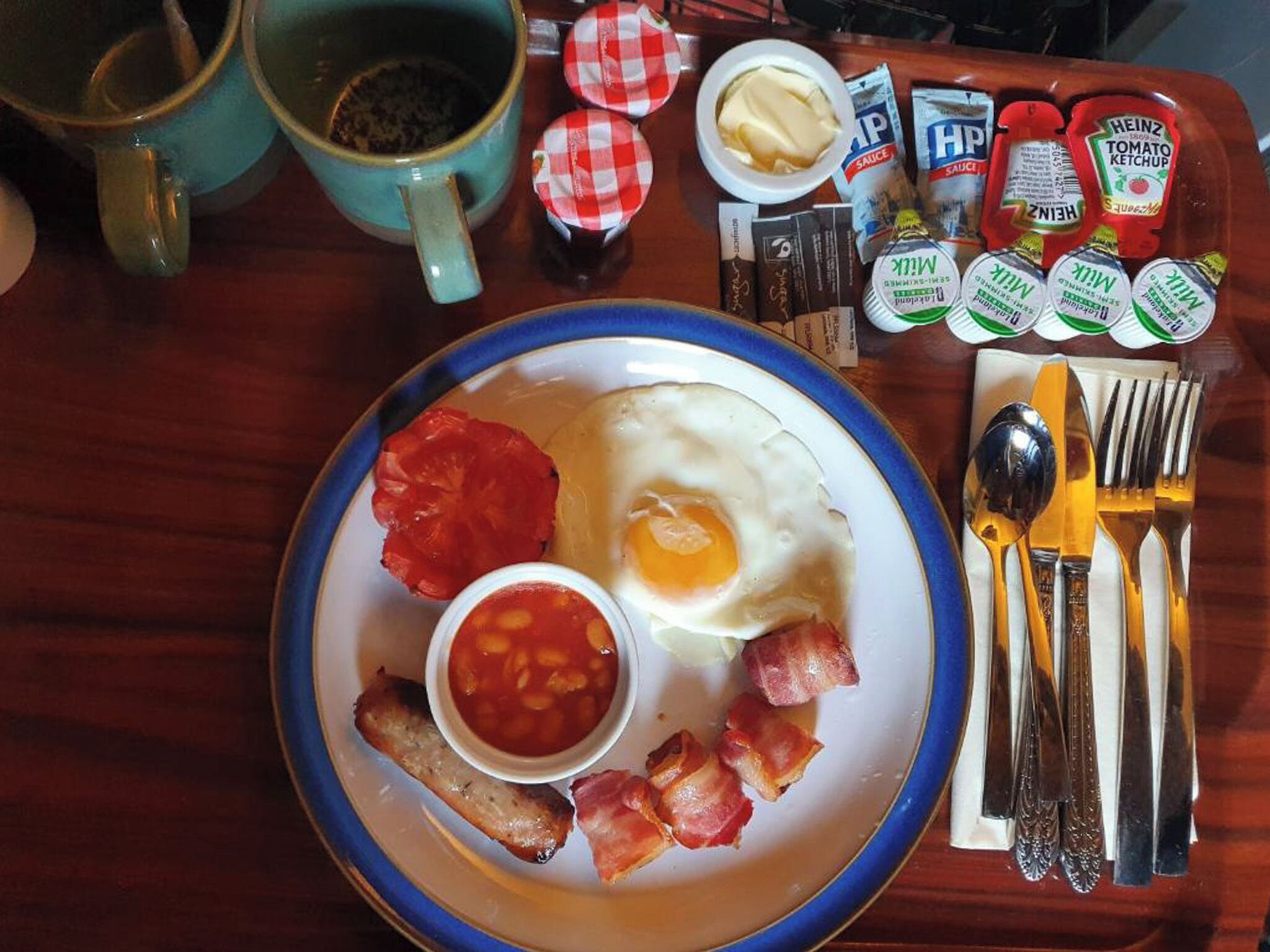 A breakfast plate with eggs, bacon, beans, and tomatoes, accompanied by condiments and utensils on a wooden table.