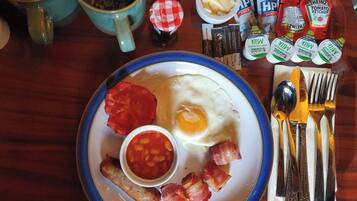 Un plato de desayuno con huevos, tocino, frijoles y tomates, acompañado de salsas y utensilios sobre una mesa de madera.