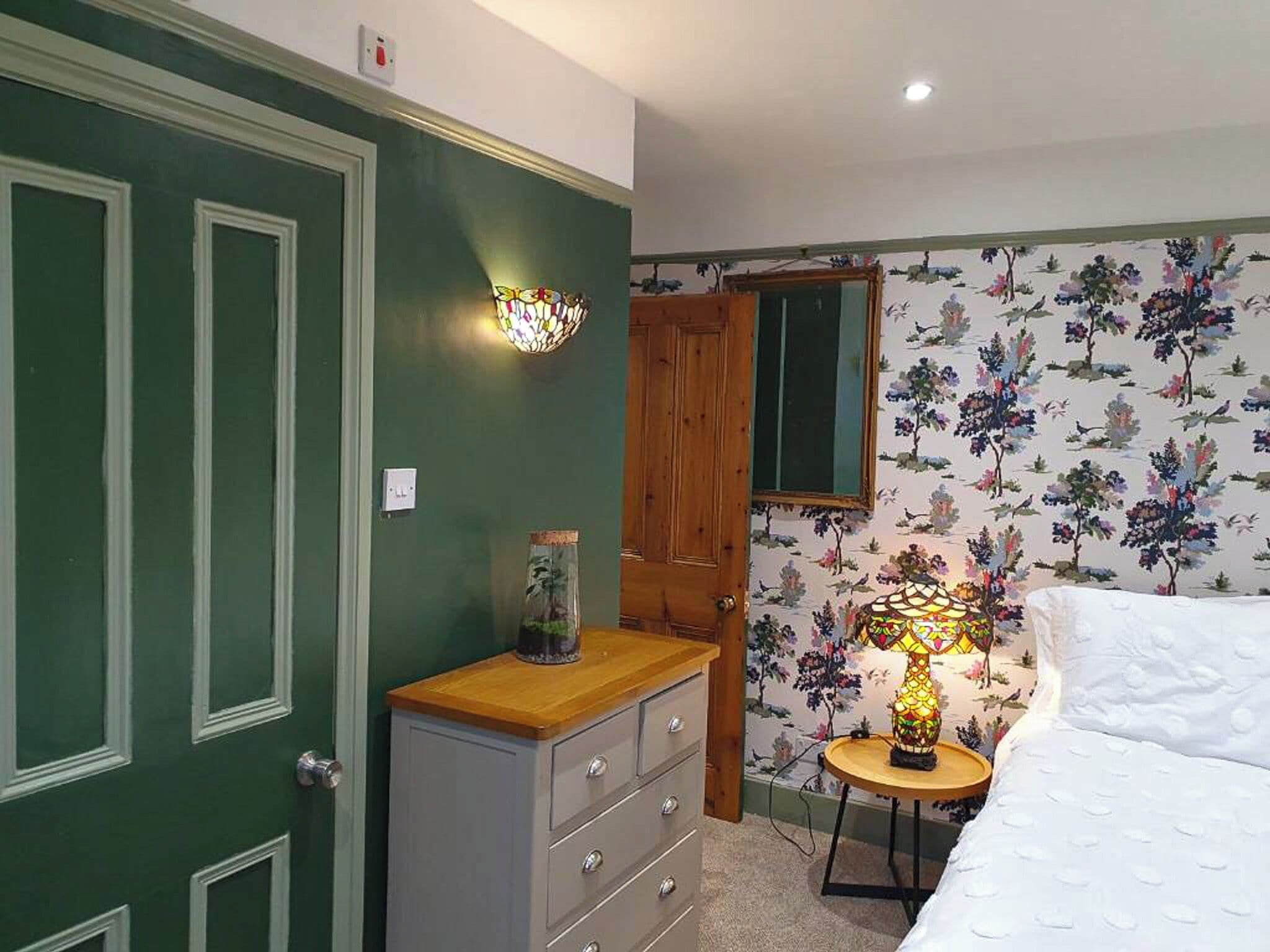 A bedroom with a green door, floral wallpaper, a wooden dresser, a bedside table with a lamp, and a bed with white bedding.