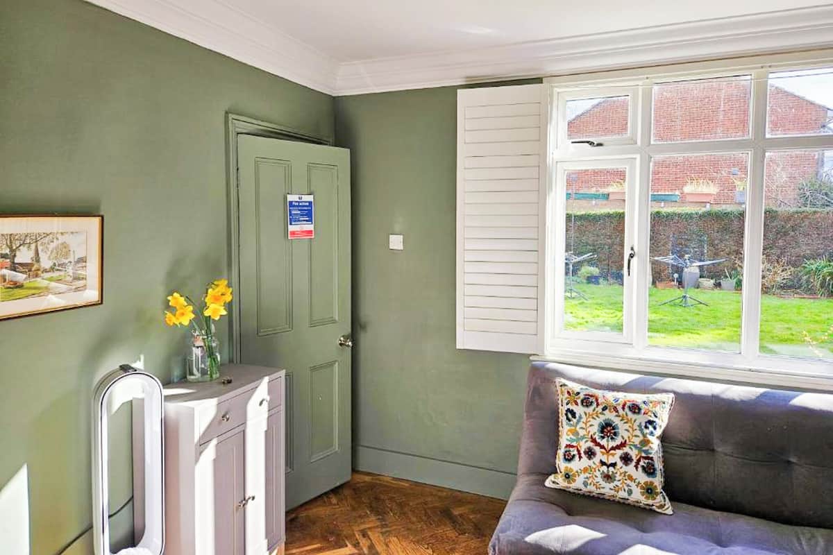 Et værelse med en grøn væg, en dør med et skilt, et vindue med skodder, en sofa med en blomstret pude og et spejl på et skab.