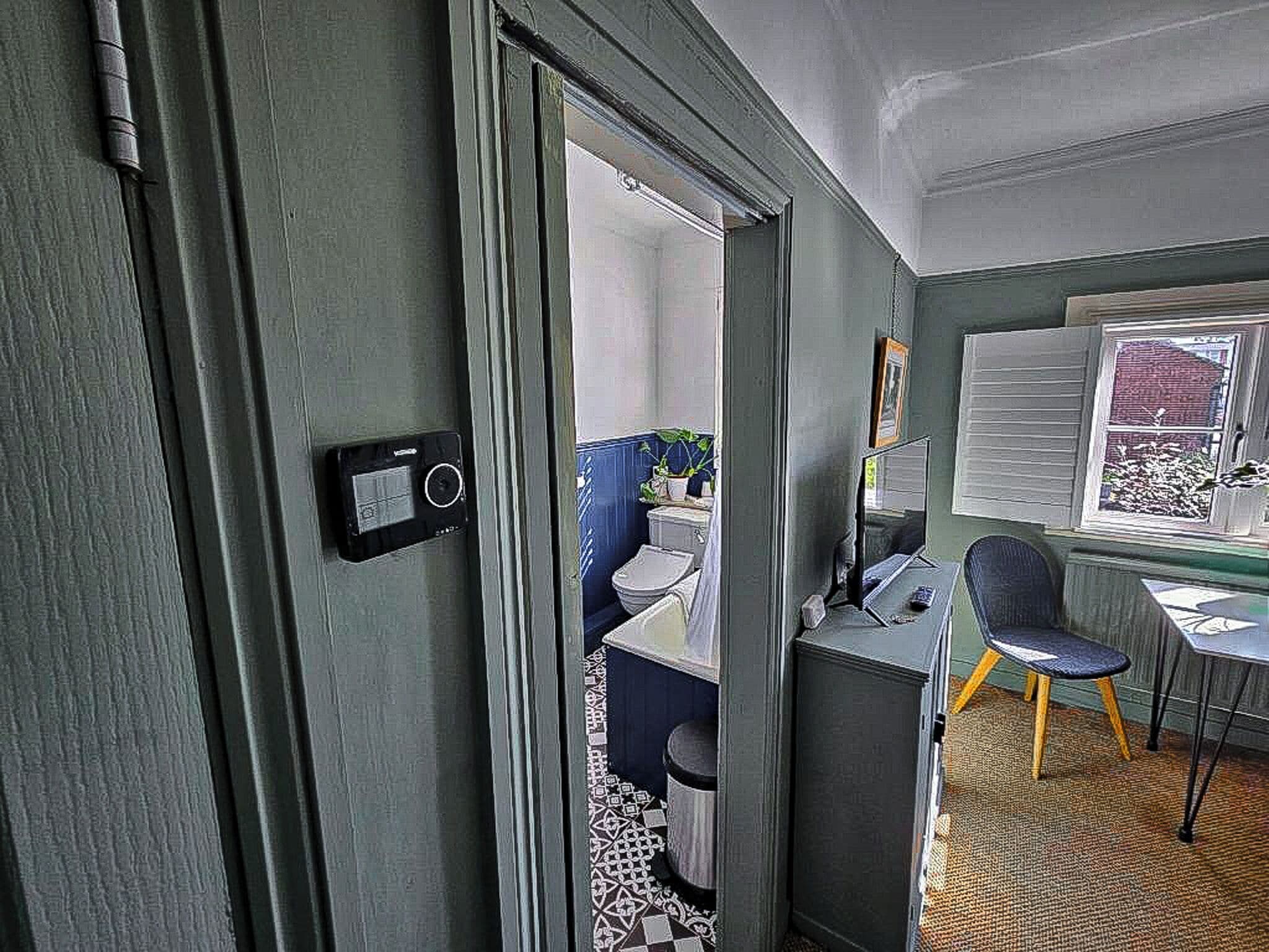 A room with a door leading to a bathroom, a desk with a chair, and a window with blinds.