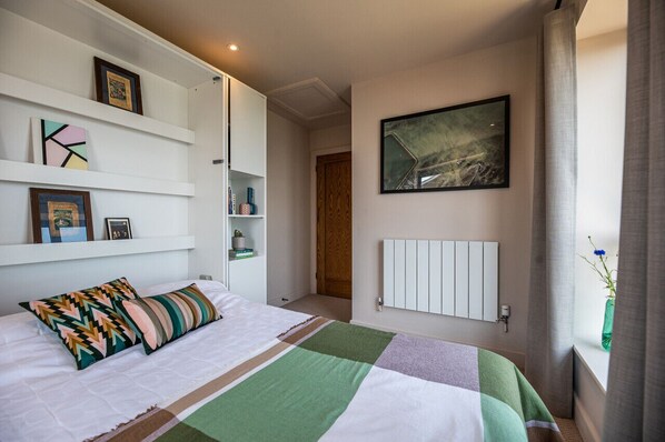 Un dormitorio con cama, un radiador, una ventana con cortinas y una pared con cuadros.