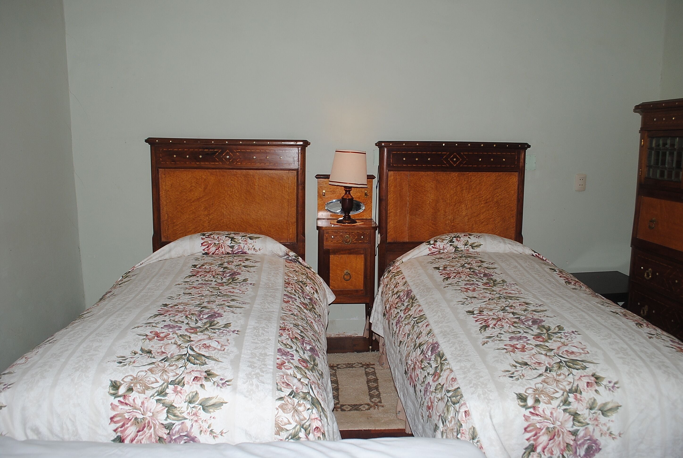 A room with two wooden beds, each with floral bedding, a small bedside table with a lamp, and a wooden dresser.