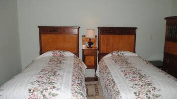 A room with two wooden beds, each with floral bedding, a small bedside table with a lamp, and a wooden dresser.