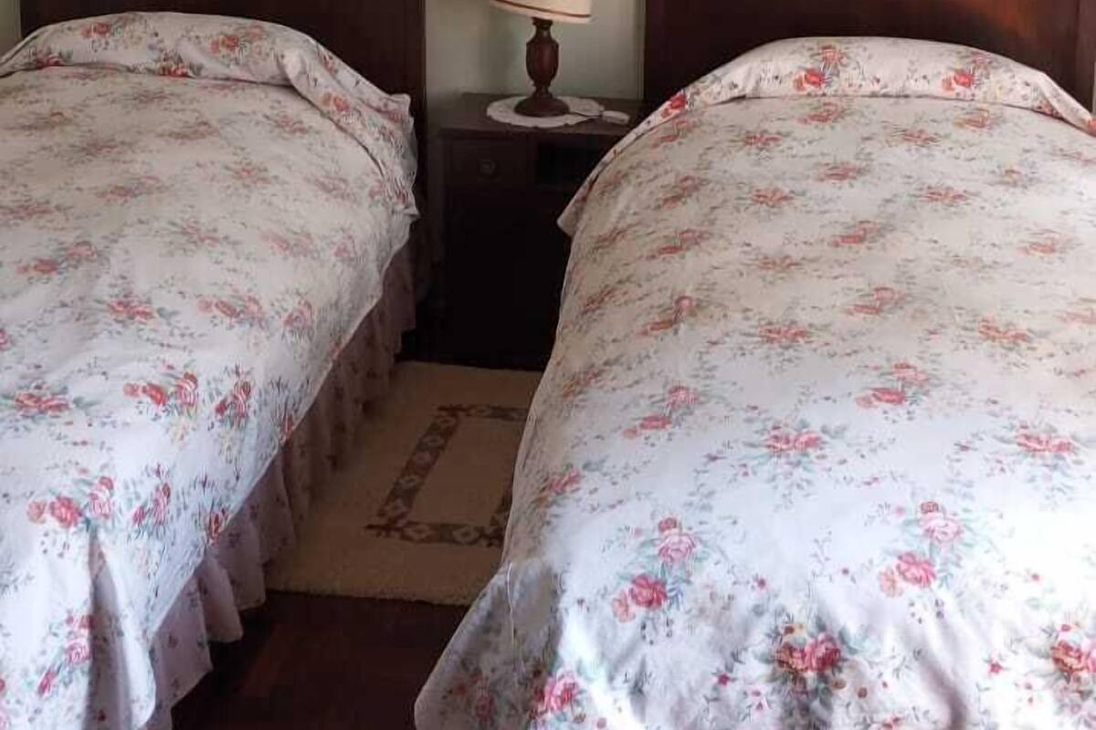 A room with two wooden beds with floral bedding, a small bedside table with a lamp, and a framed picture on the wall.