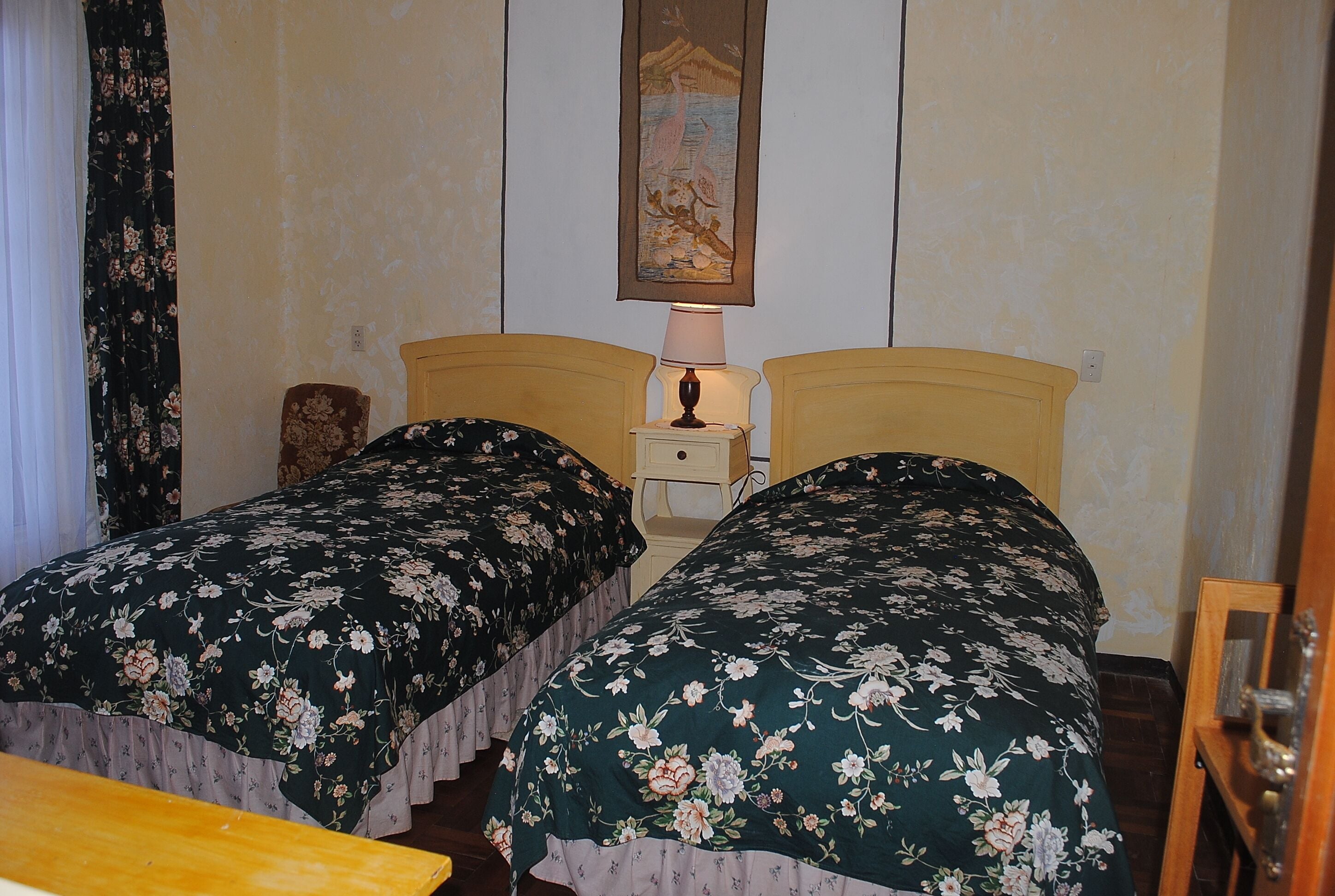 Una habitación con dos camas individuales, cada una con cubrecama floral, una mesita de noche con lámpara y una silla de madera.