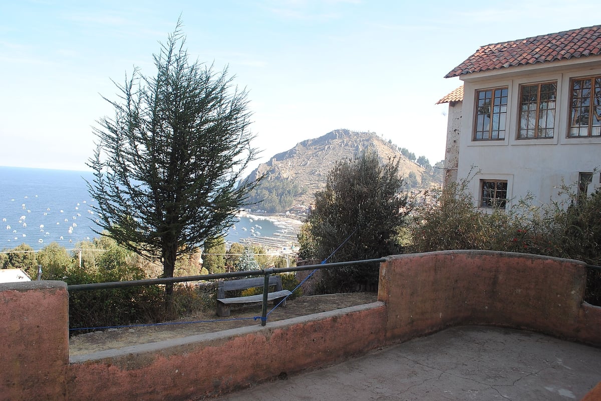 A coastal view with a mountain in the background, a bench, and a building with a tiled roof.