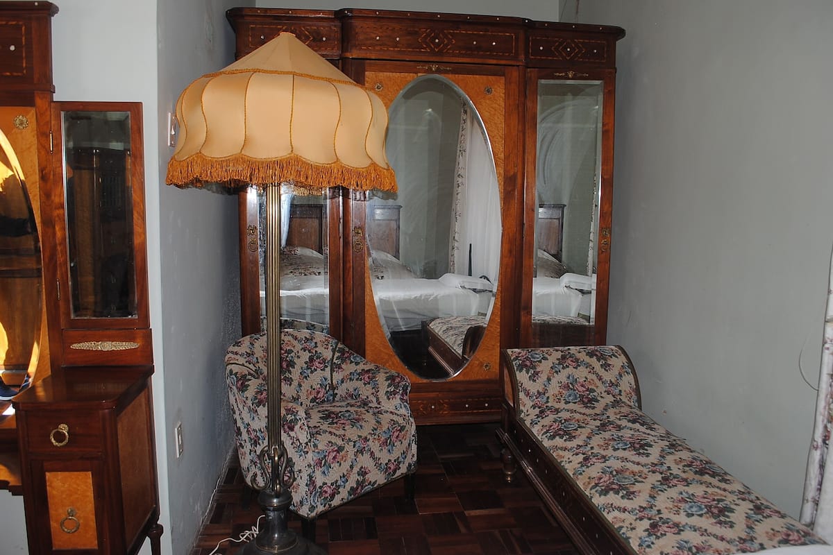 A room with a wooden wardrobe, a mirror, a floral armchair, and a small bed with a floral bedspread.