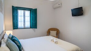 A bedroom with a bed, a window with blue shutters, a TV mounted on the wall, and a wall-mounted air conditioner. - CAM - Aguarelas de Lisboa T1 (Obidos)