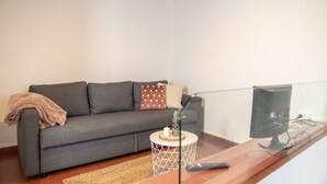 A living room with a grey sofa, a small round side table with a candle, a remote control, and a TV on a wooden stand. - CAM - Aguarelas de Lisboa T1 (Obidos)