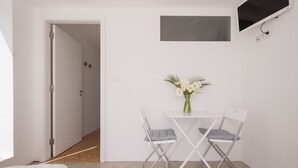 A small, modern room with a white door, a flat-screen TV mounted on the wall, a small table with a vase of flowers, and two foldable chairs. - ST Apart 01 in Atouguia da Baleia (Peniche)