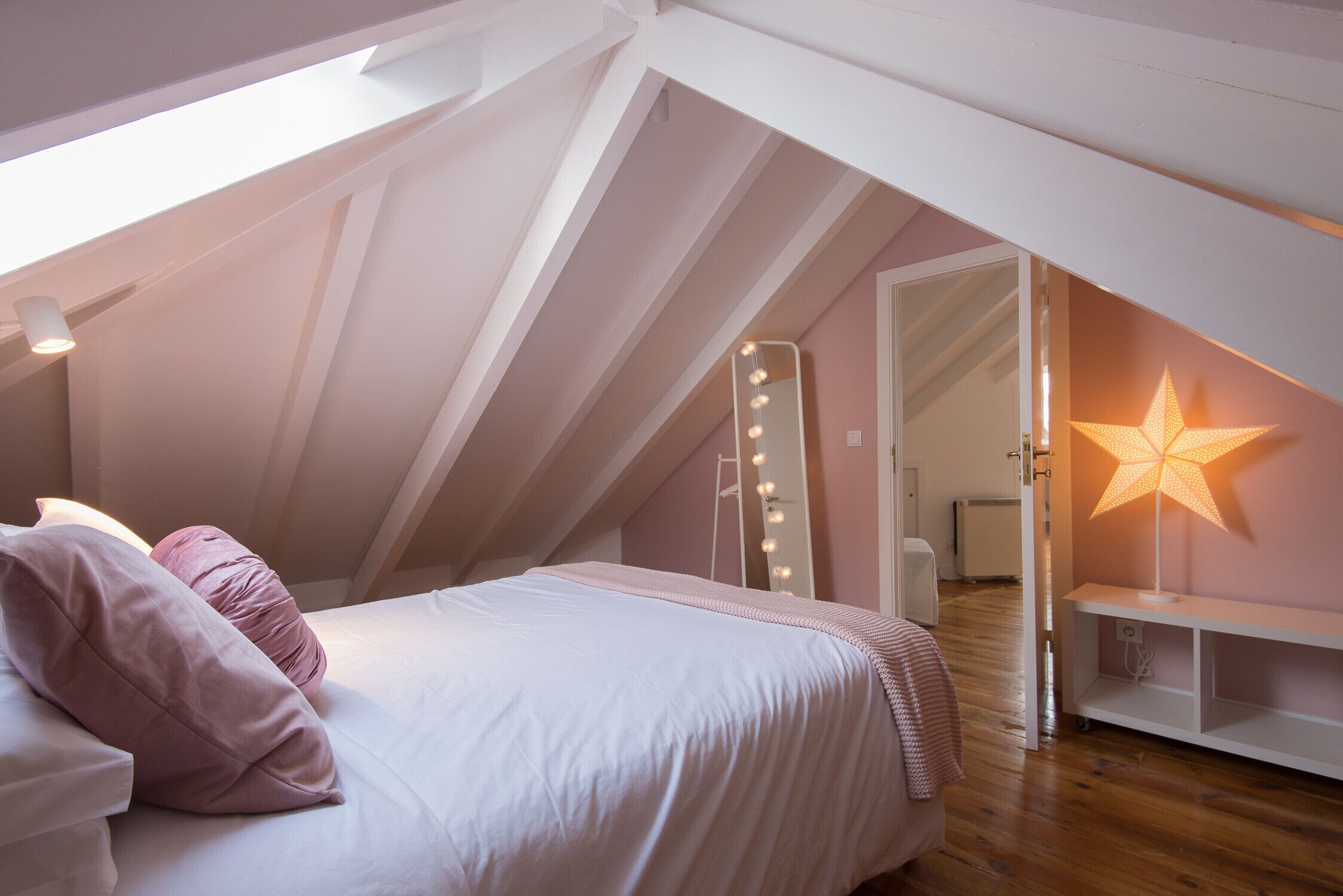 A bedroom with a sloped ceiling, a bed with white bedding and pink pillows, a star-shaped lamp, and a mirror with lights.
