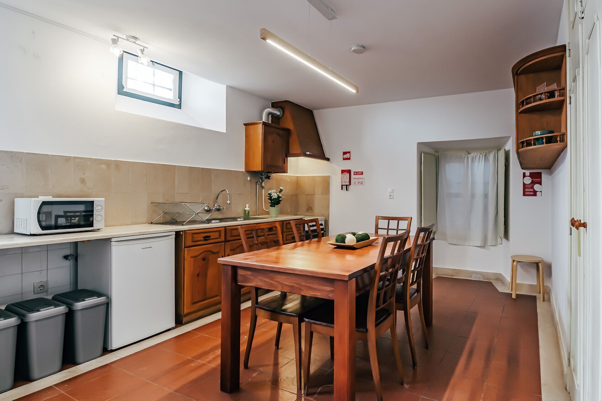 A compact kitchen with a microwave, sink, and wooden cabinets. There is a dining table with chairs and a small stool.