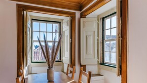 A dining area with a wooden table and chairs, a vase with dried plants, and two windows with views of buildings. - CAM - Aguarelas Francesas T2 (Obidos)