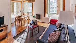 A modern living room with wooden flooring, a sofa, a dining table with chairs, a TV, and a large window. - CAM - Aguarelas Francesas T2 (Obidos)