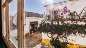A narrow street with a white building featuring a yellow stripe, a purple flower mural, and a small shop entrance. - CAM - Aguarelas Francesas T2 (Obidos)