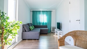 A modern living room with a grey sofa, teal curtains, a white TV stand, and a wicker chair. - Mar Alta - Supertubos (Peniche)