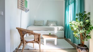 A small, well-lit room with a bed, a wicker chair, a small table with a plant, and a potted tree. - Mar Alta - Supertubos (Peniche)