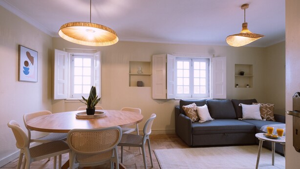 Ein modernes Wohnzimmer mit einem runden Holztisch, weißen Korbstühlen, einem grauen Sofa und zwei Pendelleuchten.