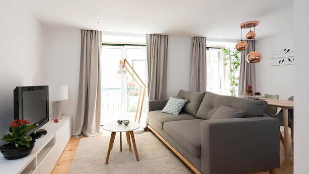 Ein modernes Wohnzimmer mit einem grauen Sofa, einem Flachbildfernseher, einem kleinen Tisch mit einer Pflanze und einem Essbereich mit Holztisch und Stühlen.