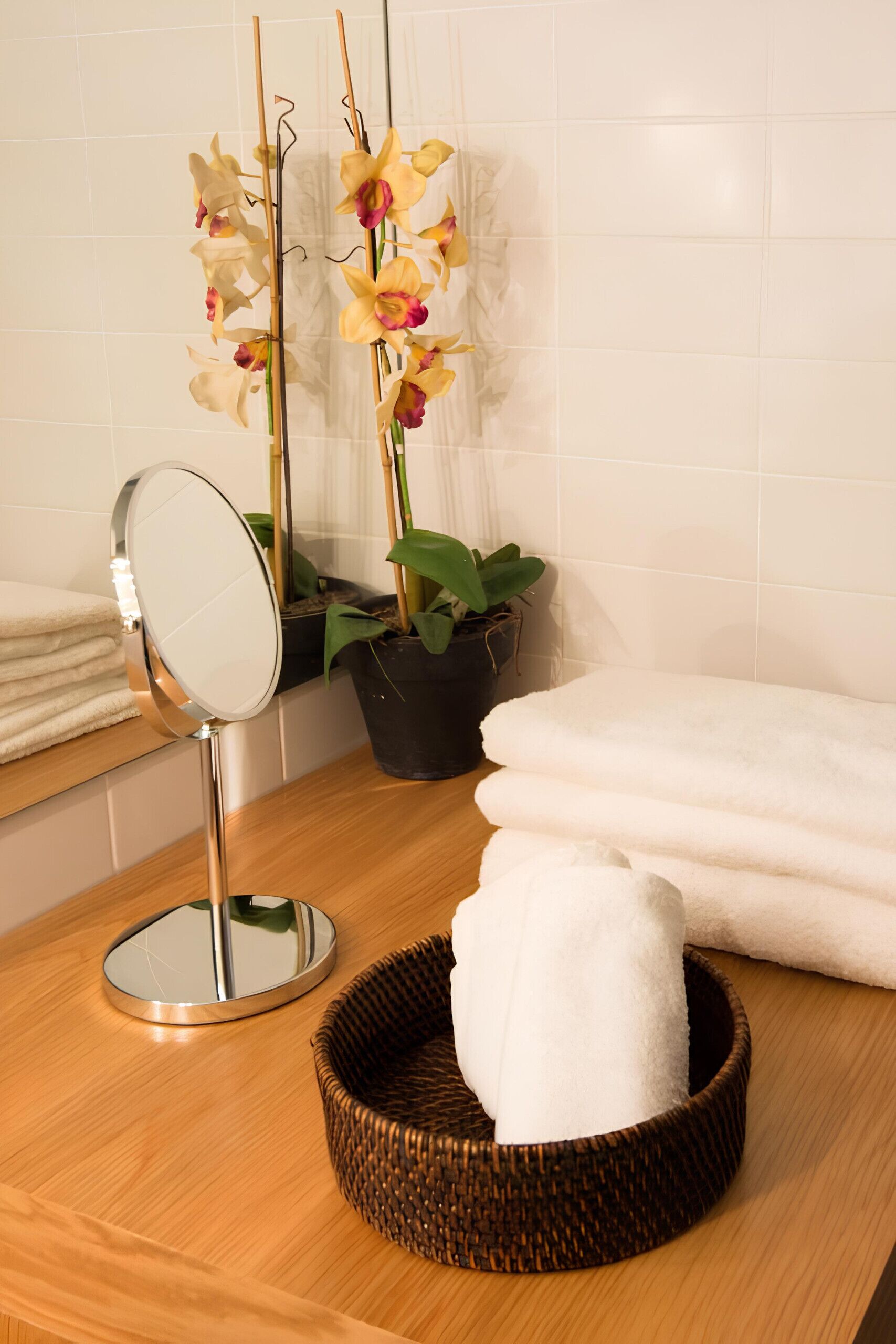 A bathroom with a wooden vanity, a mirror, a potted orchid, a stack of white towels, and a woven basket.