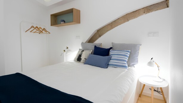 A bedroom with a bed, a bedside table with a lamp, a wooden shelf above the bed, and a coat rack.