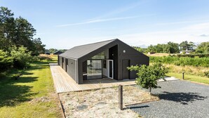 A modern, dark-colored building with a sloped roof and large glass doors, surrounded by a wooden deck and gravel path, set in a rural landscape. - Stunning home in Sæby with sauna (Sæby)
