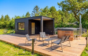 A modern outdoor deck with a wooden floor, metal chairs, a hot tub, and a small building with a dark exterior. - Stunning home in Sæby with sauna (Sæby)