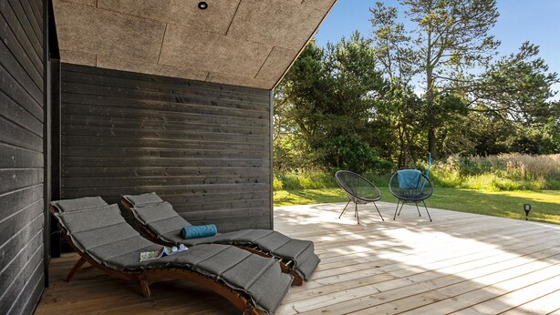 A wooden deck with two lounge chairs and a table, a wooden wall, and a garden with trees and grass. - Stunning home in Sæby with sauna (Sæby)