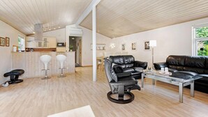 A modern living room with a leather sofa, a glass coffee table, and a black leather chair. There is a kitchen area with a microwave and a sink. The room has wooden floors and a high ceiling with exposed beams.