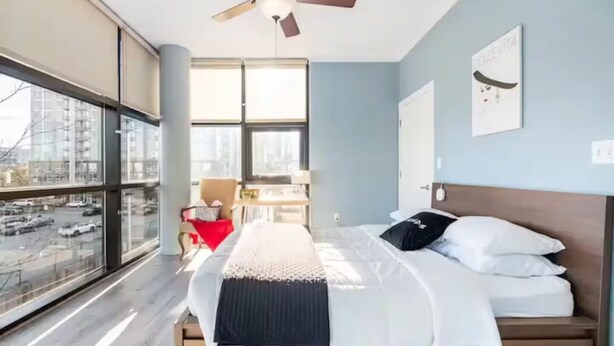 A modern hotel room with a large bed, a ceiling fan, a desk, and a view of the city through large windows. - Downtown Dream: 2-Bed Condo with Pool & Parking! (Charlotte)