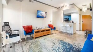 A modern living room with a ceiling fan, a flat-screen TV, a wooden cabinet, and a kitchen area with bar stools. - Downtown Dream: 2-Bed Condo with Pool & Parking! (Charlotte)