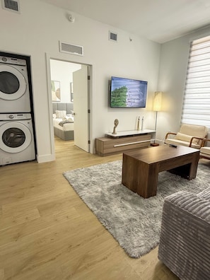 Una sala moderna con una mesa de centro de madera, una alfombra suave de color gris, un televisor montado en la pared y una zona de lavandería con lavadoras apiladas.